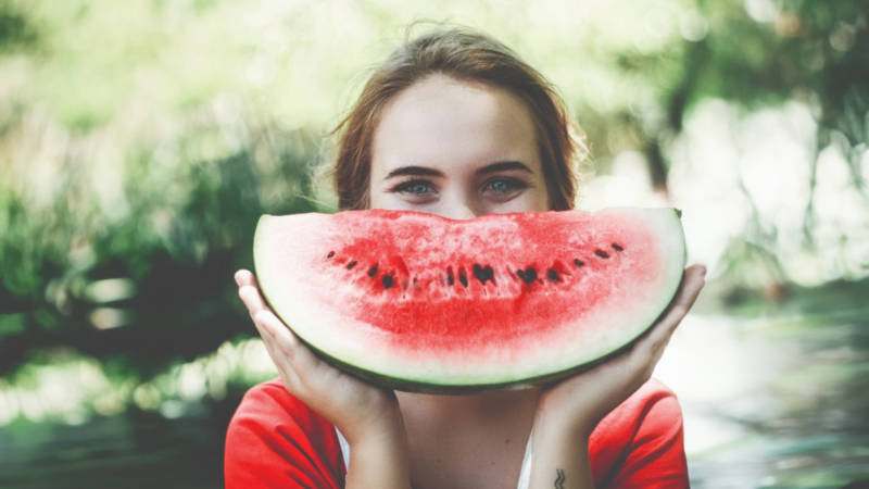 Eine Frau hält eine Scheibe Wassermelone vor ihrem Gesicht, während sie über Ernährung und mentale Gesundheit spricht.